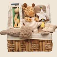 Neutral giraffe baby hamper gift set with plush toy, natural rubber teether, knitted baby blanket and newborn essentials in wicker basket nursery setting.