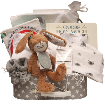 Neutral- themed baby gift set including a plush bunny toy, knitted baby booties, 'Guess How Much I Love you' baby blanket, and star patterned suitcase. 
