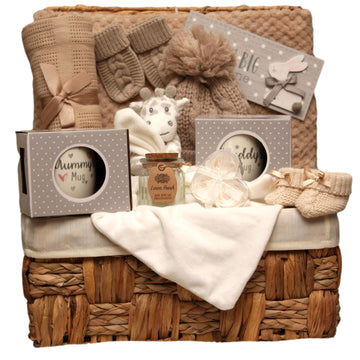 Wicker baby gift hamper filled with beige knitted blankets, baby socks, a soft giraffe toy, mugs and pampering items in neutral tones. 